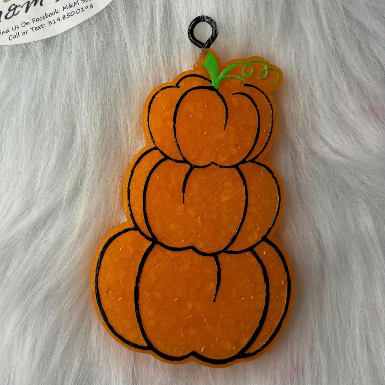 A plastic air freshener in the shape of a stacked pumpkin placed on a white furry surface with a label that reads 'M&M Scents & Apparel'.