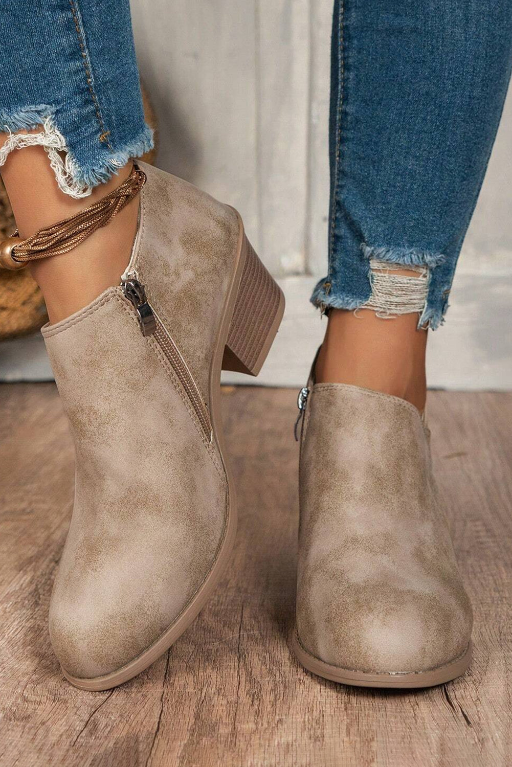 A pair of beige suede ankle boots with a lace-up closure and a block heel, shown against a backdrop of a person's lower legs and jeans.