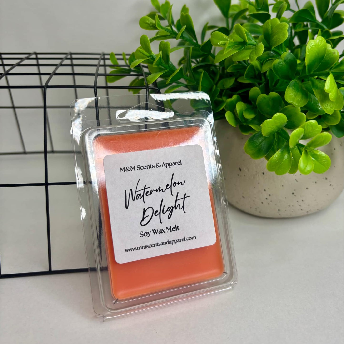 A clear container with an orange wax melt inside, labeled 'Watermelon Delight Soy Wax Melts'. The container is placed on a white surface with a plant in the background.