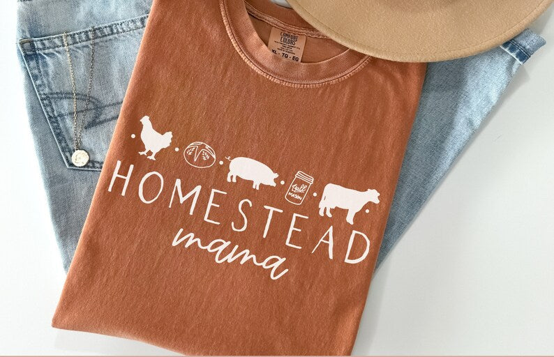 A rust-brown graphic t-shirt laid flat on top of light-wash blue jeans, with a tan hat partly visible at the top. The shirt has a white printed design reading “HOMESTEAD mama” with small farm-themed silhouettes.