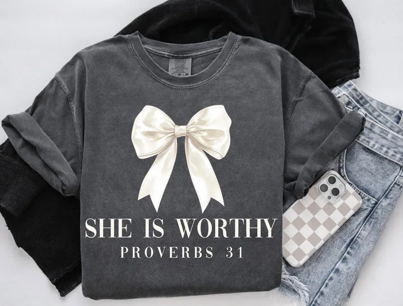 Dark gray t-shirt with a white bow and text 'She is Worthy Proverbs 31' on a light background.