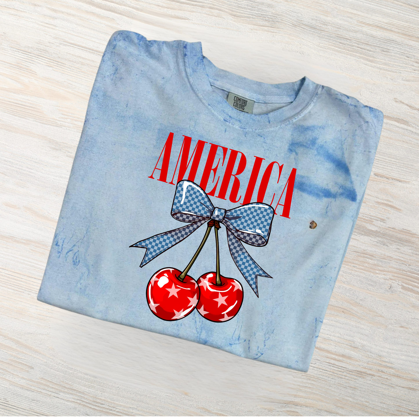 Light blue t-shirt with 'AMERICA' and cherry design on a wooden surface