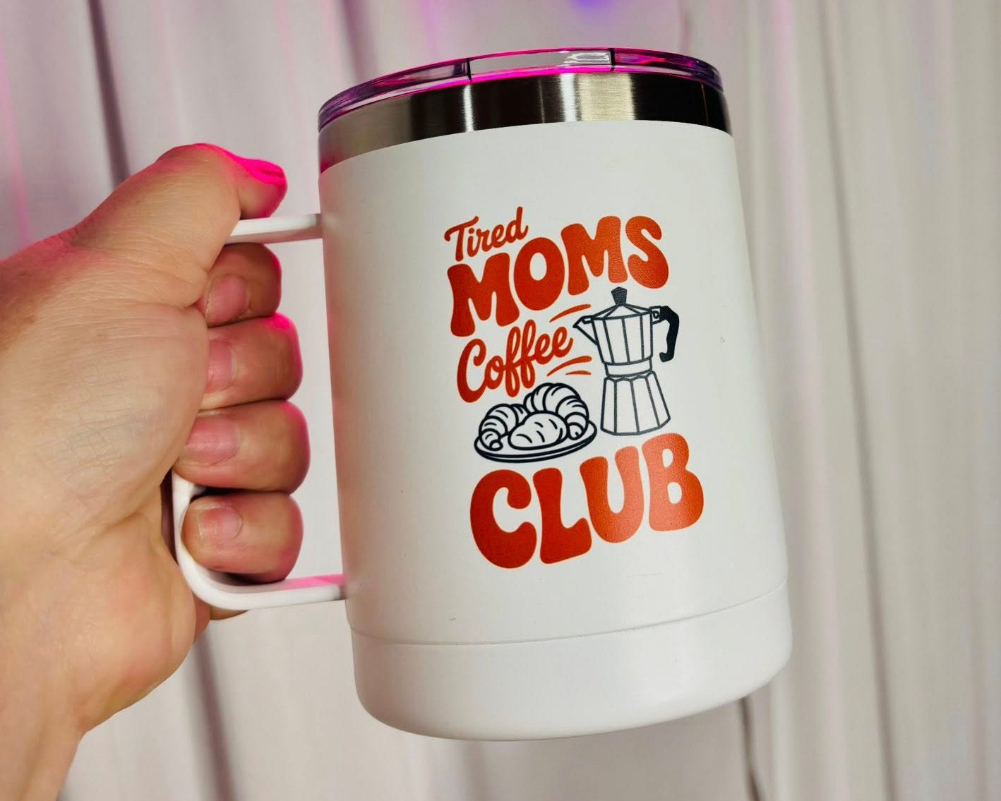 White mug with 'Tired Moms Coffee Club' design held by a hand, neon sign in the background.