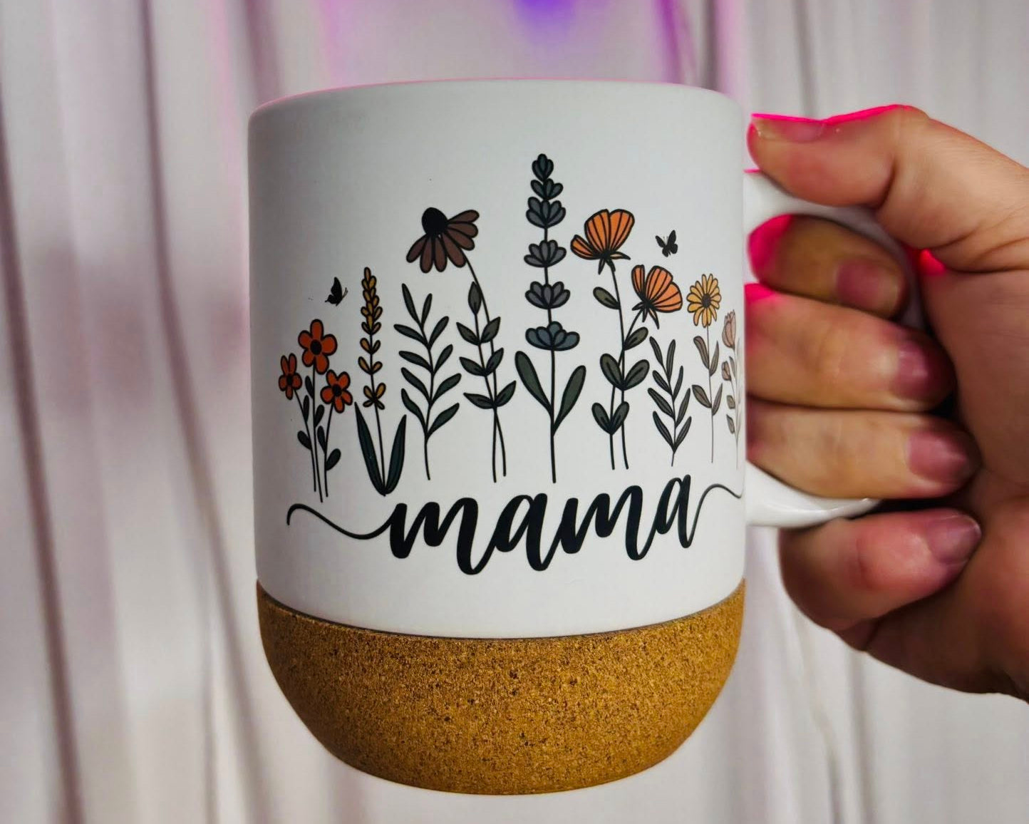 Mug with floral design and 'mama' text held by a hand in front of a neon sign.
