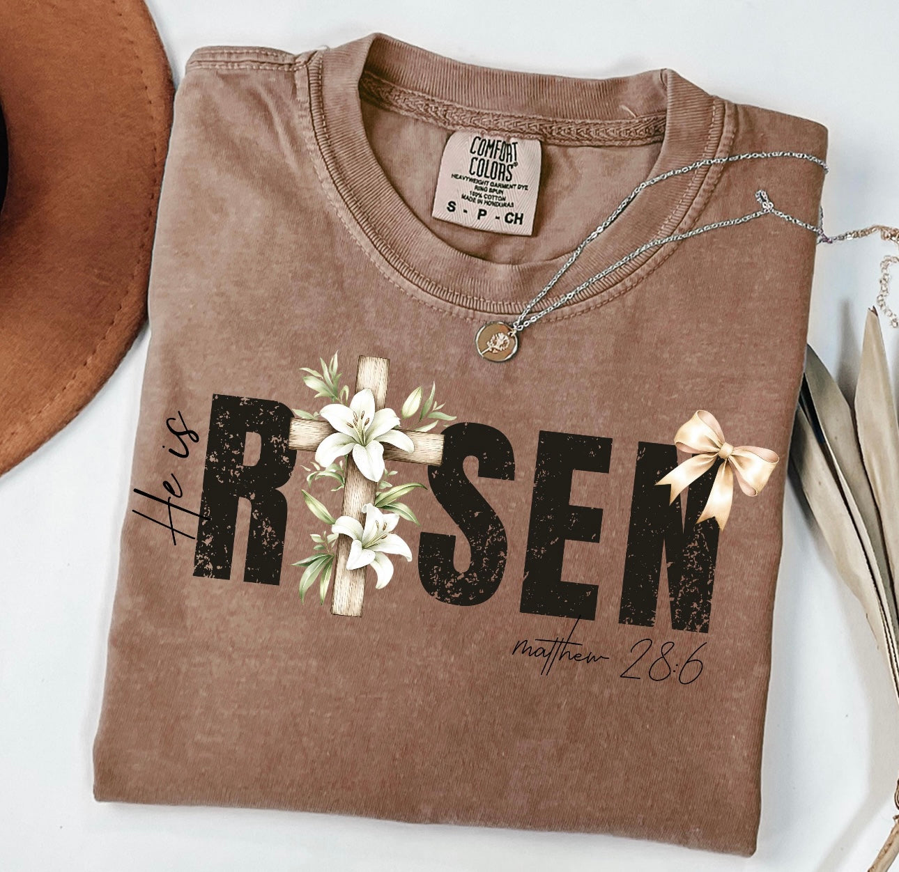 Brown t-shirt with 'He is Risen' text and cross design on a white background
