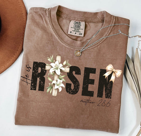 Brown t-shirt with 'He is Risen' text and cross design on a white background