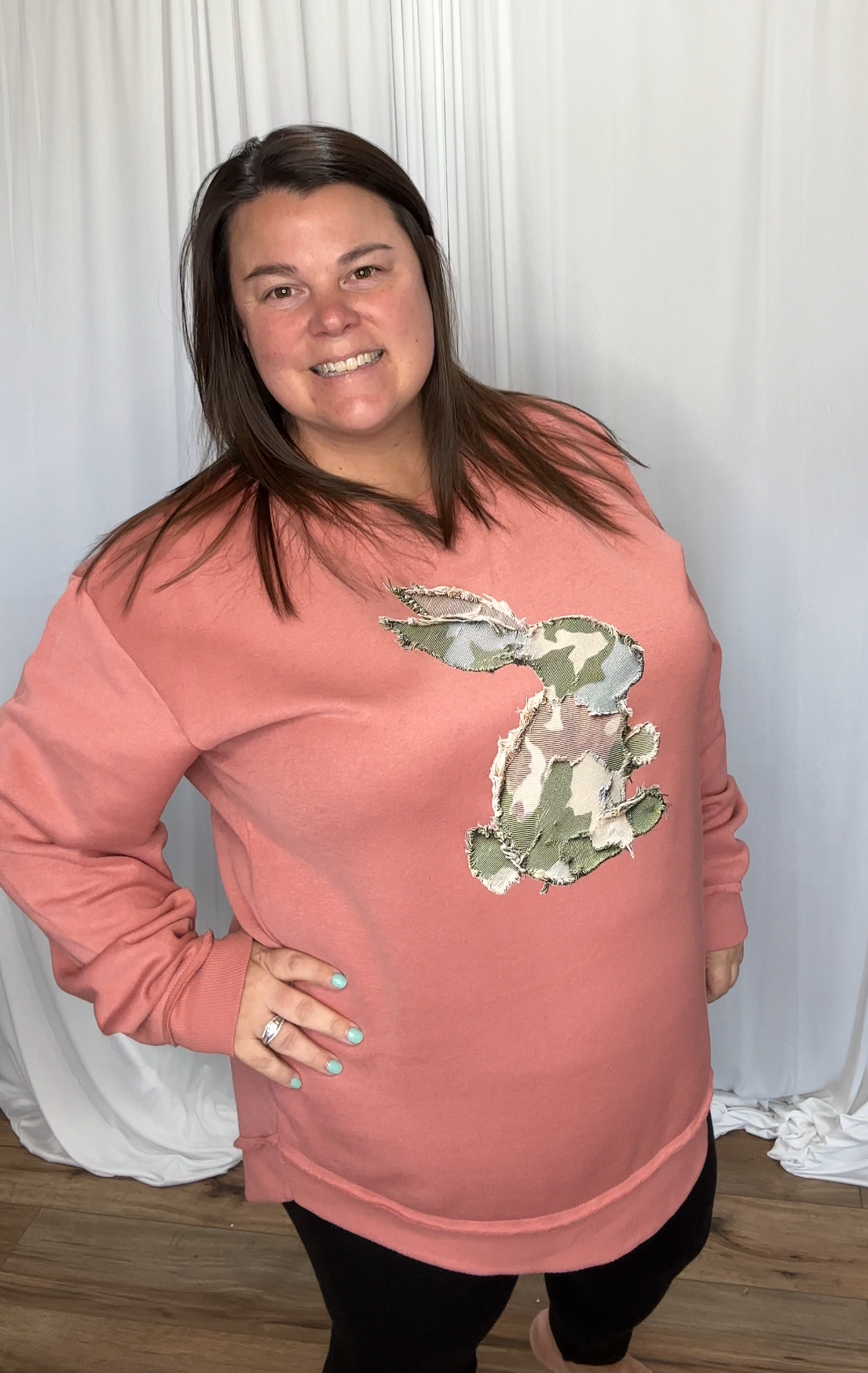 Woman wearing a pink sweatshirt with a green graphic design against a white curtain background.