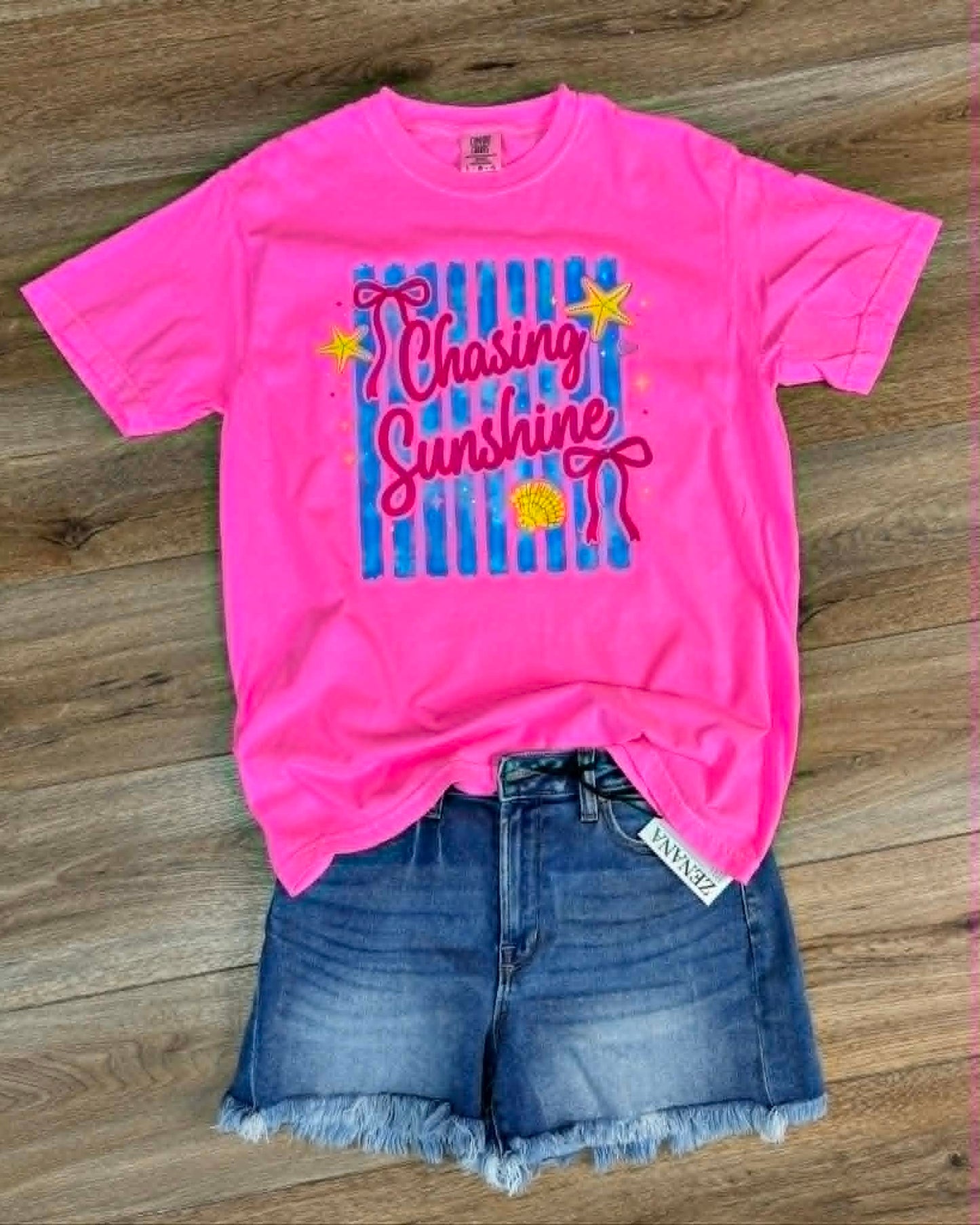 Pink t-shirt with 'Chasing Sunshine' design and denim shorts on a wooden surface
