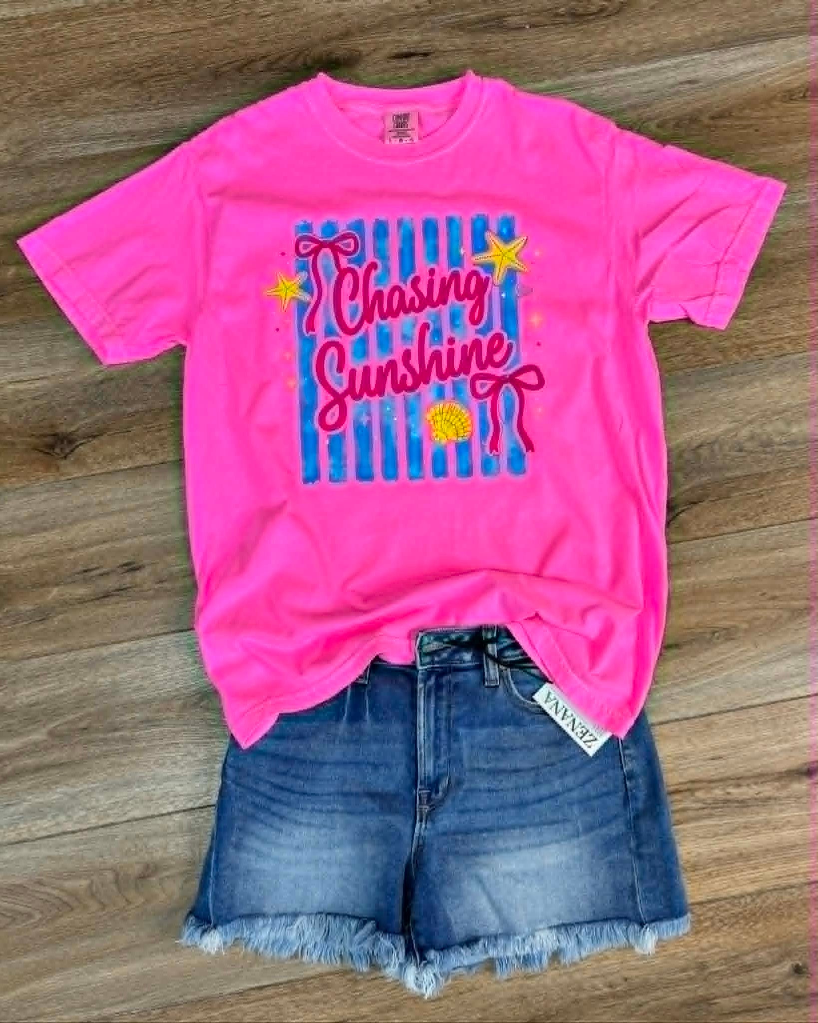 Pink t-shirt with 'Chasing Sunshine' design and denim shorts on a wooden surface