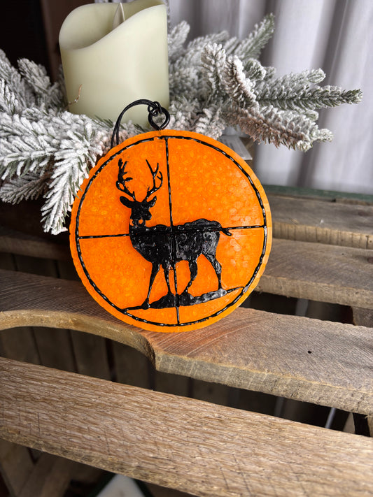 Decorative ornament with deer silhouette on an orange background, placed on a wooden surface with flocked branches.