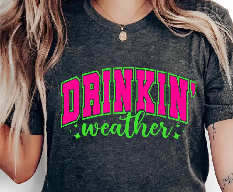 Person wearing a dark gray t-shirt with 'DRINKIN' weather' text in pink and green.
