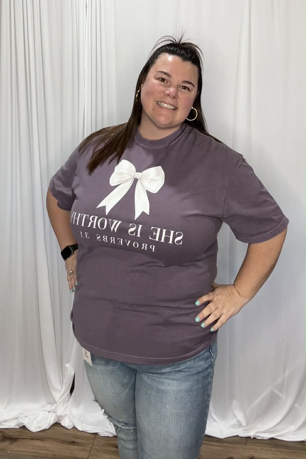 Woman wearing a purple t-shirt with text and a white bow graphic against a white curtain background