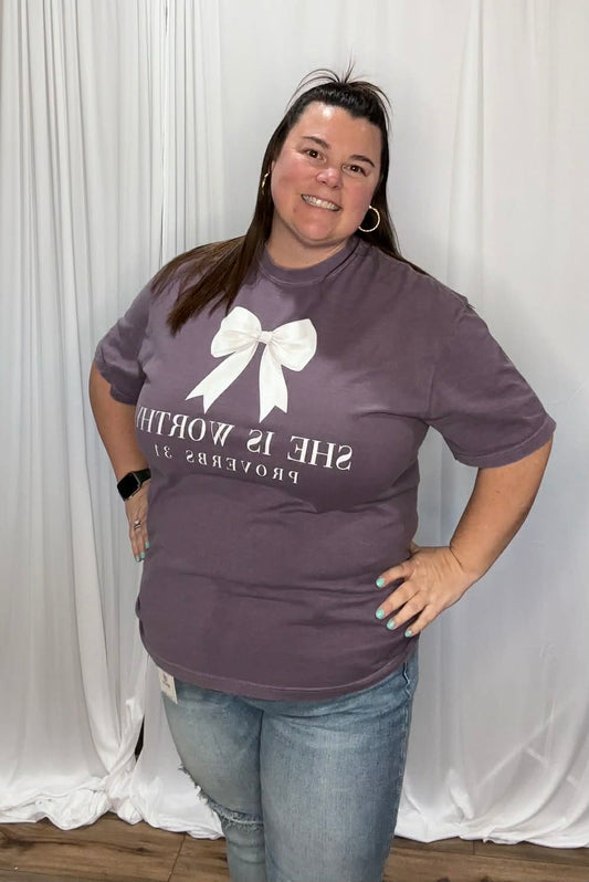 Woman wearing a purple t-shirt with text and a white bow graphic against a white curtain background