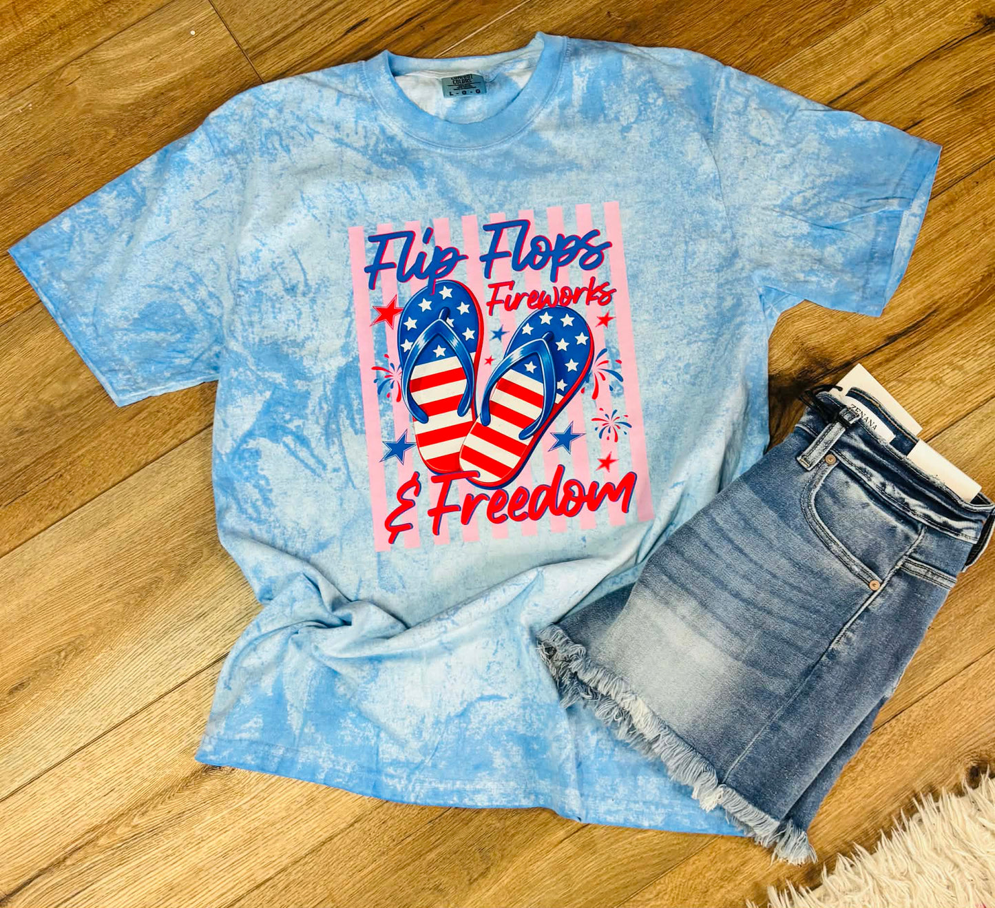 Blue tie-dye t-shirt with 'Flip Flops, Fireworks & Freedom' graphic on a wooden floor.