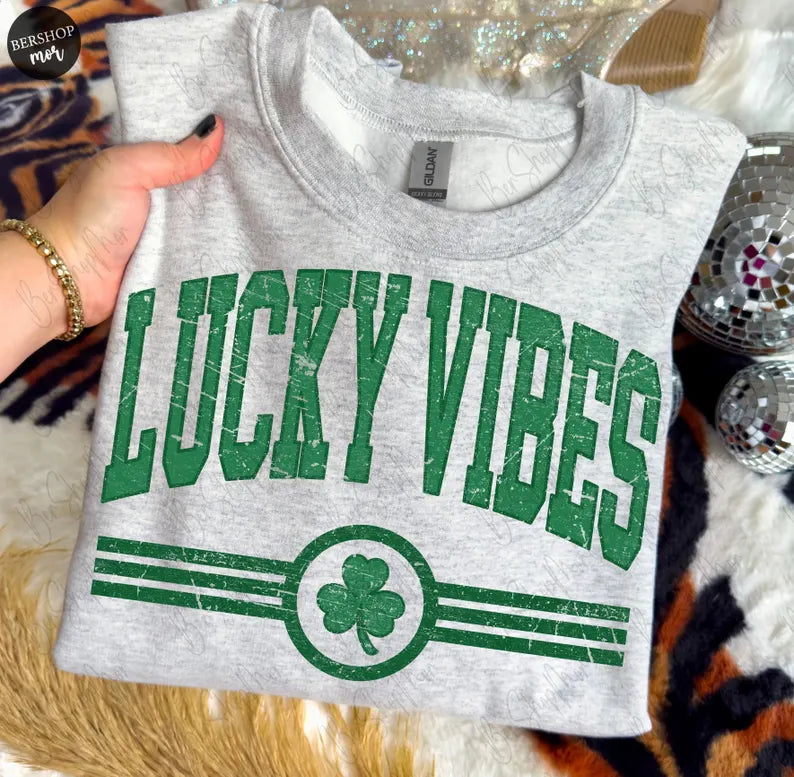 Gray tank top with 'Lucky Vibes' text and shamrock design held by a hand.