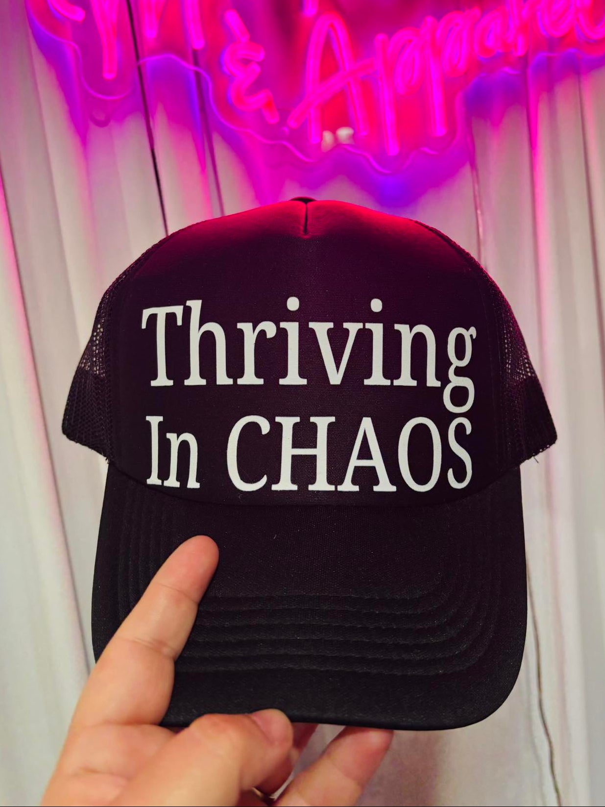 Black cap with 'Thriving In CHAOS' text held in front of a neon sign.