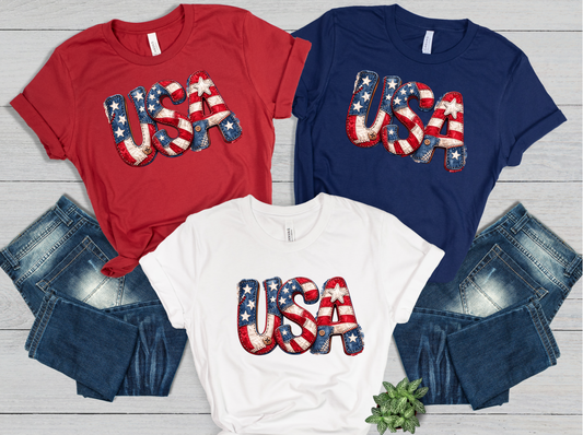 Three t-shirts with 'USA' design on a wooden surface with jeans.